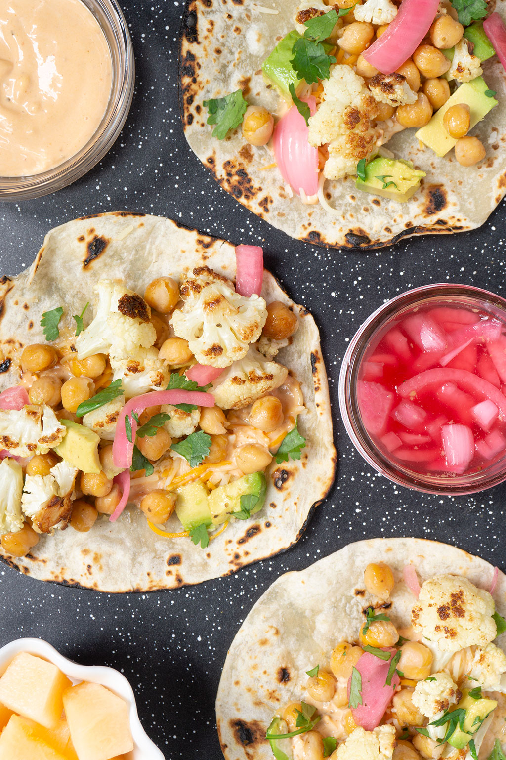 vertical image of cauliflower tacos on tray, bowl of fruit on side